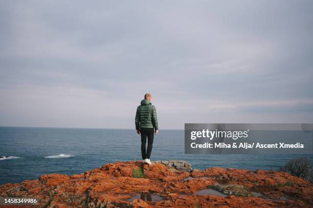 young man with camera explores rocky outcrop above sea - terrasse panoramique photos et images de collection