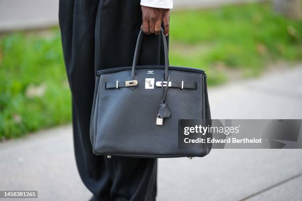 Guest wears a white shirt leather zipper jacket, black flowing suit pants, a black shiny leather Birkin handbag from Hermes, outside Hermes, during...