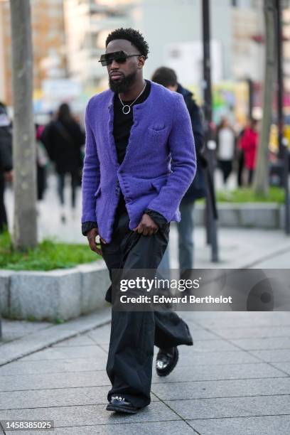 Guest wears black futurist sunglasses, a black t-shirt, a silver circle pendant necklace, a purple tweed buttoned blazer jacket, black cargo pants,...