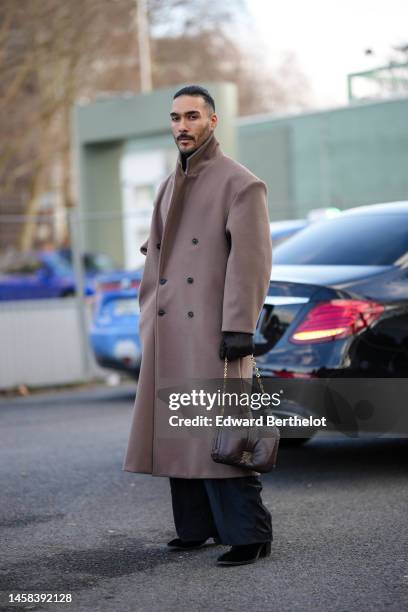 Guest wears a black turtleneck pullover, a brown beige long wool coat, a brown shiny leather puffy handbag from Loewe, black wide legs pants, black...