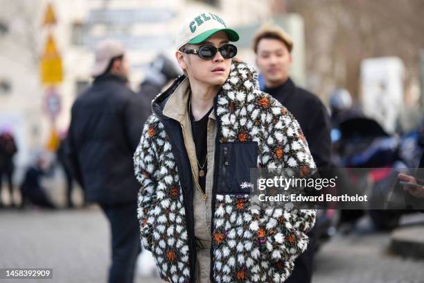 Guest wears a beige with green embroidered ripped logo cap from Celine, black sunglasses from Celine, a beige zipper jacket, a brown / white / orange...