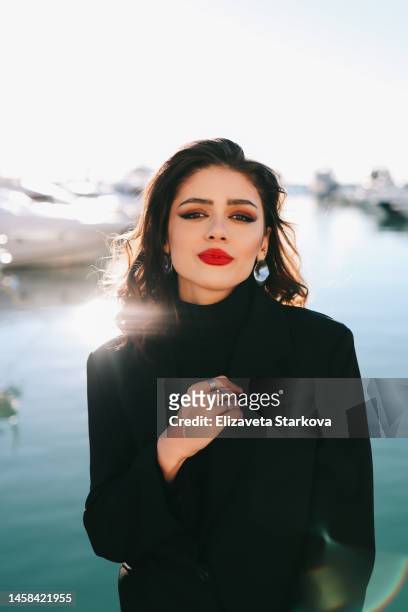 a large portrait of a young brunette woman with bright makeup and red lips with jewelry and earrings in her ears in a black stylish and fashionable suit looks into the camera against the background of the blue sea and yachts outdoor - red lipstick stock pictures, royalty-free photos & images
