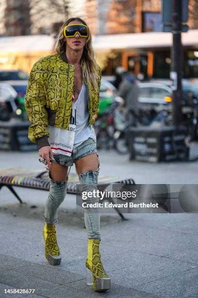 Guest wears a yellow solar ski mask, a white V-neck tank-top, a yellow with black snake print pattern shiny leather zipper coat, a white with black...