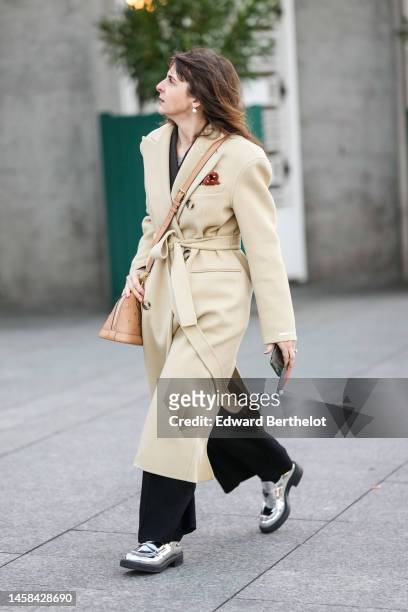 Guest wears earrings, a black blazer jacket, a beige wool long belted coat, a beige shiny leather with gold embroidered LV monogram pattern shoulder...