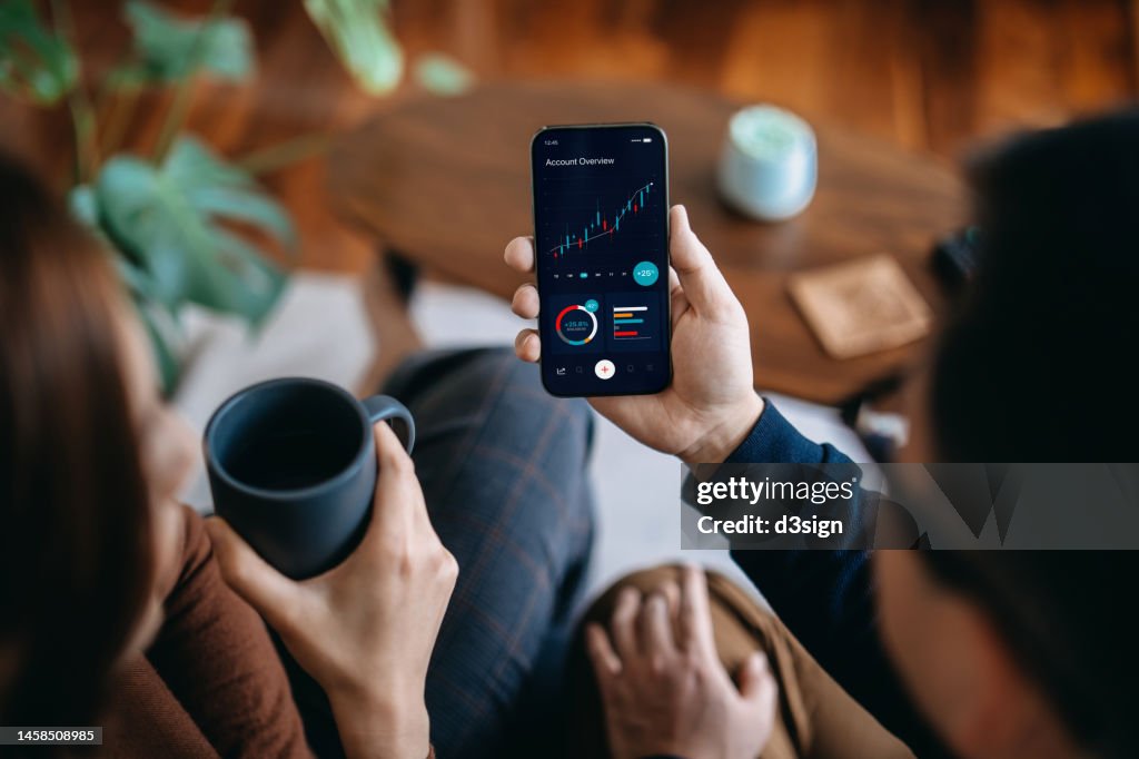 Young Asian couple managing finance and investment online, analyzing stock market trades with mobile app on smartphone. Making financial plans. Banking and finance, investment, financial trading, mobile banking concept
