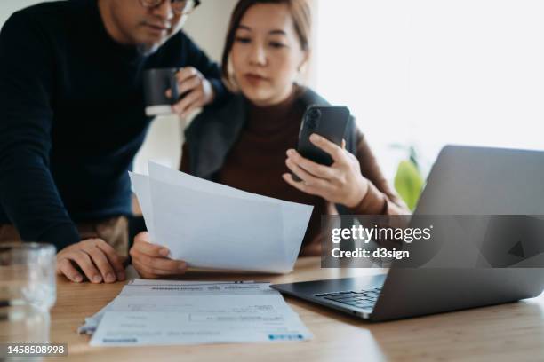 asian couple sitting at dining table, working on their finances with laptop together. making financial plans. manage stock trades. checking financial bills and calculating expenses, paying bills online, manage budget and preparing tax documents at home - fees concept stock-fotos und bilder