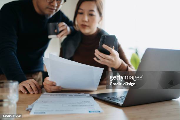 asian couple sitting at dining table, working on their finances with laptop together. making financial plans. manage stock trades. checking financial bills and calculating expenses, paying bills online, manage budget and preparing tax documents at home - self employed photos et images de collection