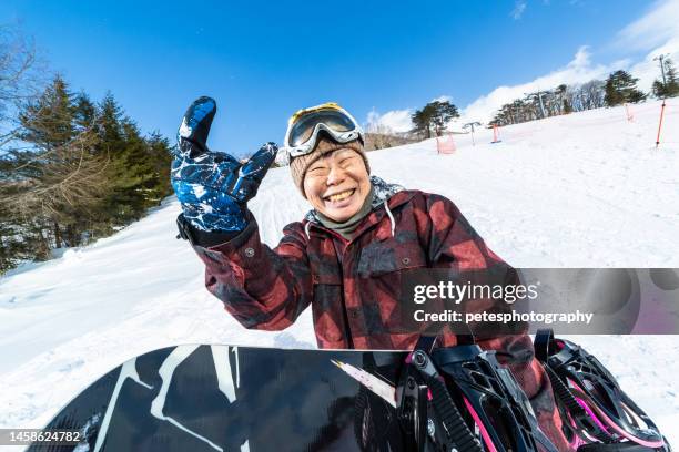 a cheerful young at heart senior snowboarder woman with her snowboard giving a "devil horns" hand gesture - radical snowboarding stock pictures, royalty-free photos & images