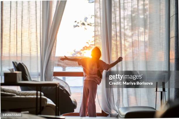 the girl opens the curtains of a large panoramic window and illuminates the room with warm sunset light. - home golden hour stock pictures, royalty-free photos & images