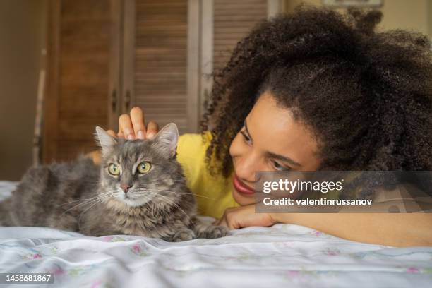 jeune femme afro à la maison, caressant son chat domestique, allongée sur le lit - cats on the bed photos et images de collection