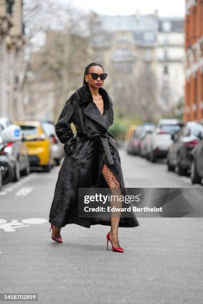 Emilie Joseph wears black sunglasses, a black long fur belted coat from the Frankieshop, black fishnet / mesh tights, dark burgundy shiny leather...