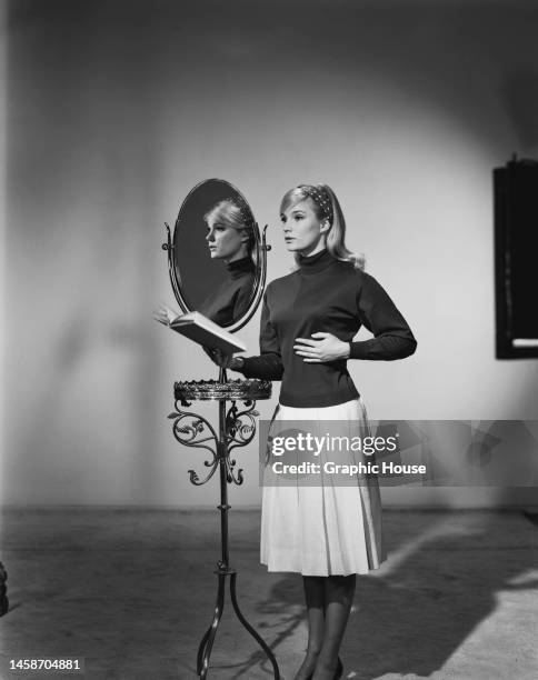 American actress Yvette Mimieux wearing a black turtleneck sweater and a white skirt, her right hand holding an open book, her left hand pressed...