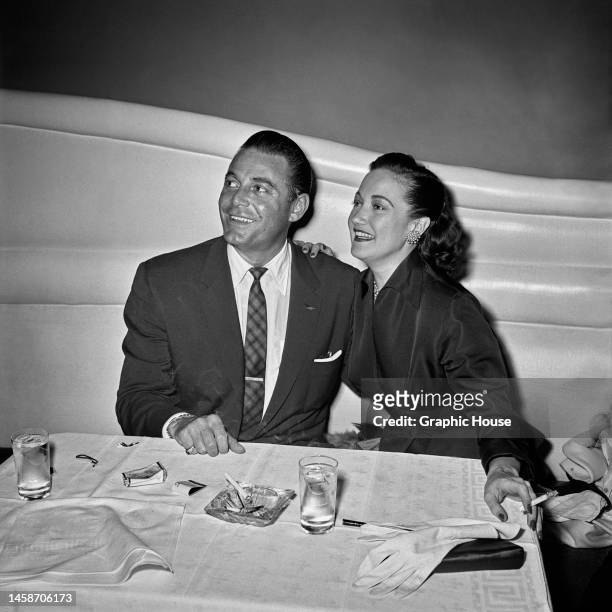 American actor Jon Hall , holding a lit cigarette as the pair sit at a dining table on which is an ashtray and a cigarette along with two drinking...