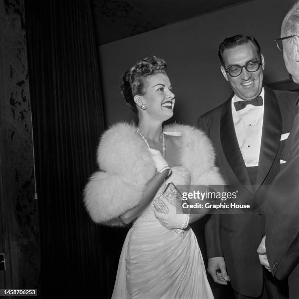 American actress and singer Gale Storm , wearing a white fur evening wrap over a white evening gown, holding clutch purse in her white-gloved hand,...