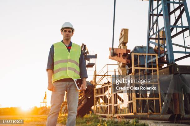 male engineer in hardhat standing against nodding donkey, pump oil jack with tablet - oil production platform stock pictures, royalty-free photos & images