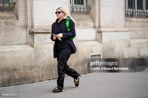 Guest wears black sunglasses, a black hoodie sweater, a black striped print pattern blazer jacket, a green shiny sequined shoulder bag, black shiny...