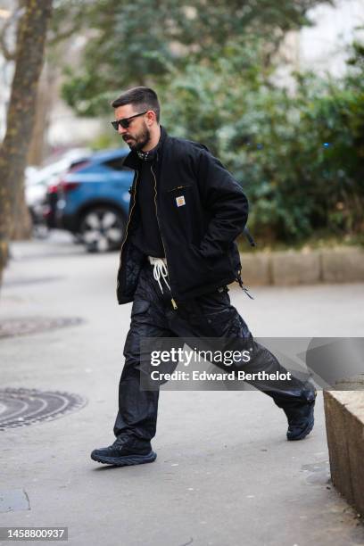 Guest wears black sunglasses, a black with white print pattern scarf, a black pullover, a black zipper oversized coat from Carhartt, black shiny...