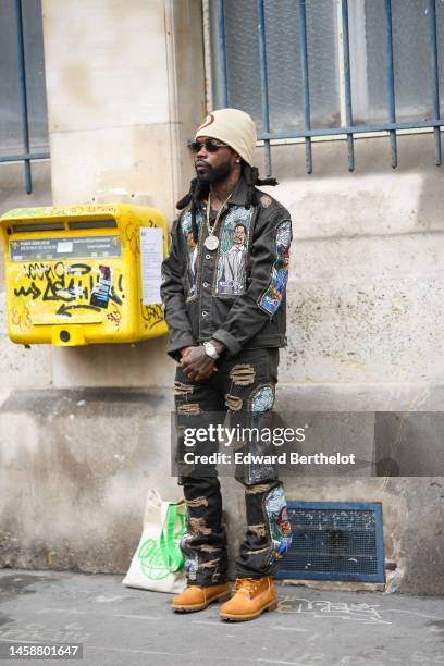 Guest wears a beige and camel print pattern wool beanie, black sunglasses, diamonds earrings, a gold large pendant necklace, a gray t-shirt, a black...