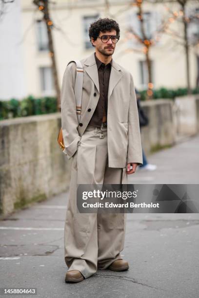 Guest wears black glasses, a brown shirt, a beige oversized blazer jacket, matching beige suit pants, a camel shiny leather shoulder bag, brown shiny...