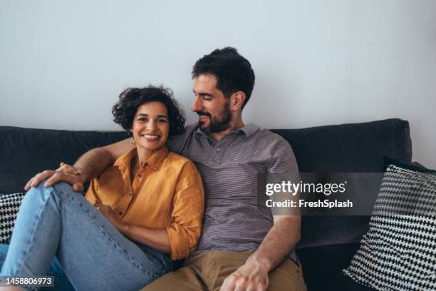 una hermosa pareja feliz mirando a la cámara mientras está sentada en el acogedor sofá y disfrutando de pasar su tiempo libre juntos - women in transparent clothing fotografías e imágenes de stock