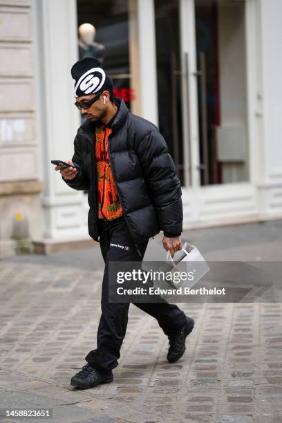 Guest wears a black and white print pattern wool beanie, black sunglasses, a black high neck puffer jacket, black pants, an orange with burgundy and...