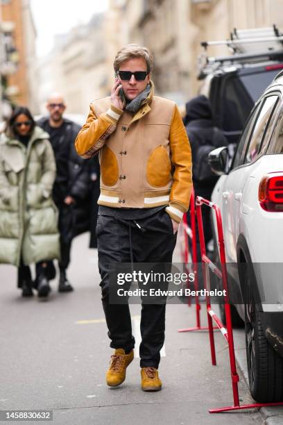 Guest wears black sunglasses, a gray wool scarf, a beige leather and camel sleeves coat, black large pants, yellow suede sneakers from Adidas ,...