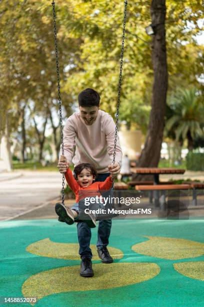 father playing with his son on the swing in the park - family in the park stock pictures, royalty-free photos & images