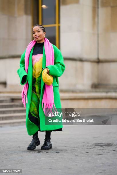 Ellie Delphine wears gold earrings, a black with pink / yellow / green print pattern high neck / long flowing dress, a neon green wool long oversized...