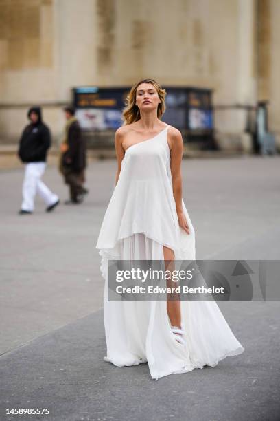 Guest wears a white asymmetric shoulder / long tulle dress, white matte leather puffy strappy heels sandals, outside Georges Hobeika, during Paris...