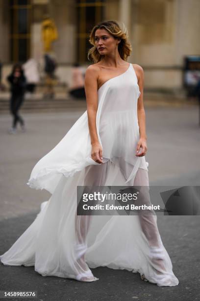 Guest wears a white asymmetric shoulder / long tulle dress, white matte leather puffy strappy heels sandals, outside Georges Hobeika, during Paris...