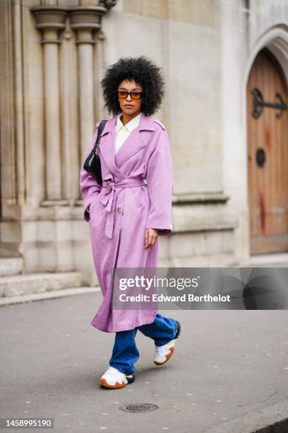 Guest wears black and orange sunglasses, a white shirt, a pale purple linen belted long coat, a black shiny leather shoulder bag, blue denim flared...