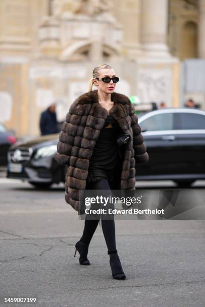 Guest wears black sunglasses from Versace, earrings, a dark brown fur oversized long jacket, a black cut-out chest short dress, black tights, black...