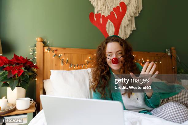 woman wearing antler and clown's nose waving on video call through laptop in bedroom - clowns nose stock pictures, royalty-free photos & images