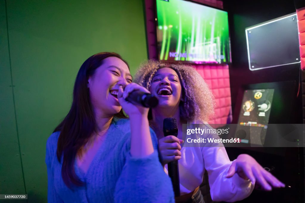 Happy young friends singing karaoke in microphone at amusement arcade
