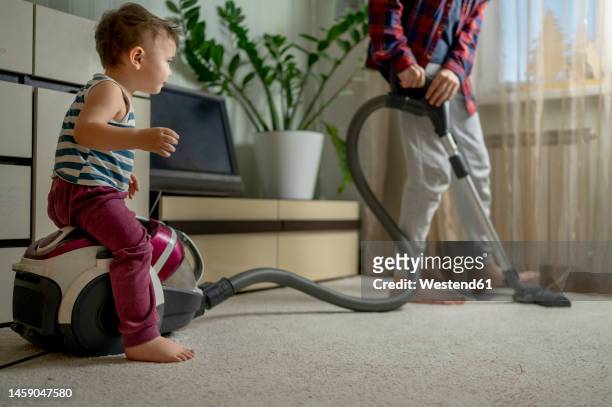 boy cleaning carpet with brother sitting on vacuum cleaner at home - low section stock-fotos und bilder
