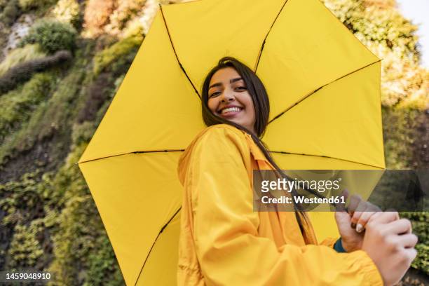 smiling young woman holding yellow umbrella in front of vertical garden - holding umbrella stock pictures, royalty-free photos & images