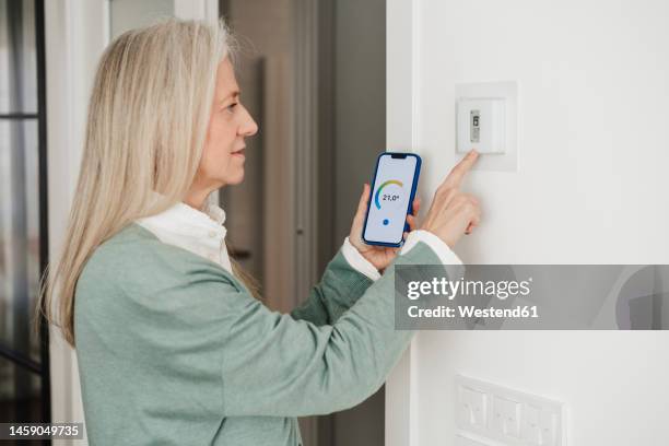 mature woman adjusting thermostat with smart phone at home - adjusting stock pictures, royalty-free photos & images