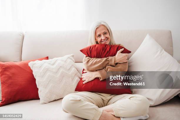 happy mature woman with cushion sitting on sofa at home - cushion imagens e fotografias de stock
