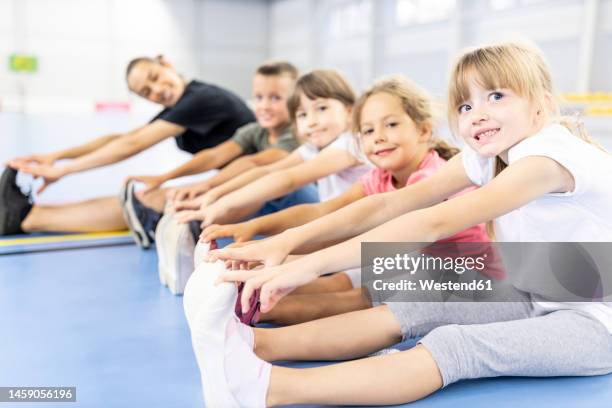 smiling elementary students doing exercise sitting side by side with teacher at school sports court - portrait of school children and female teacher in field stock pictures, royalty-free photos & images