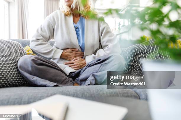 woman having stomach ache sitting on sofa at home - bauchweh stock-fotos und bilder