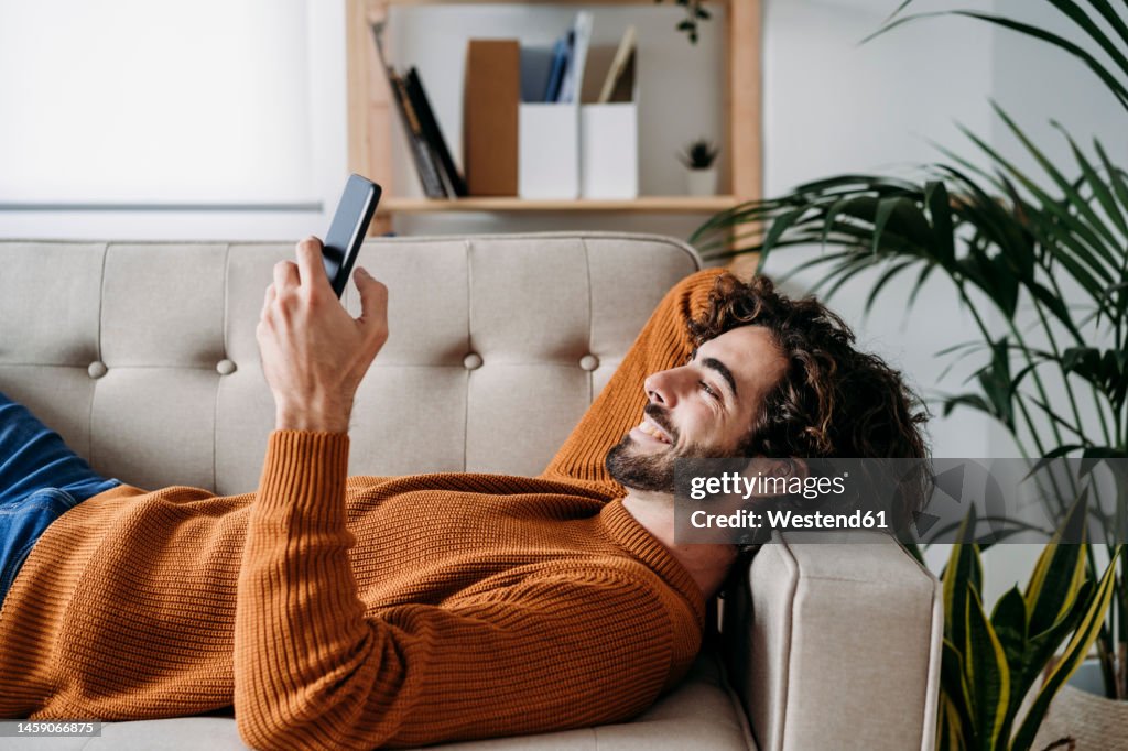 Happy young man using smart phone relaxing on sofa at home