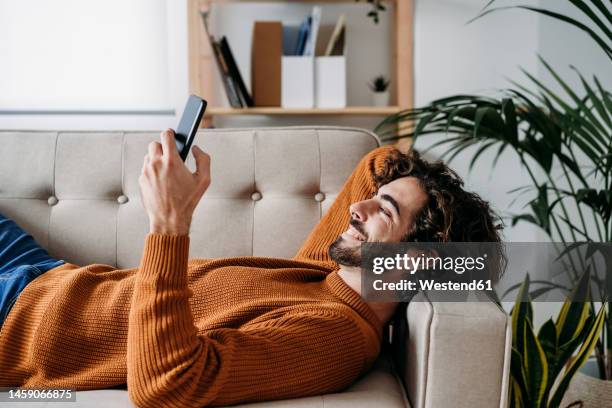 happy young man using smart phone relaxing on sofa at home - homens imagens e fotografias de stock