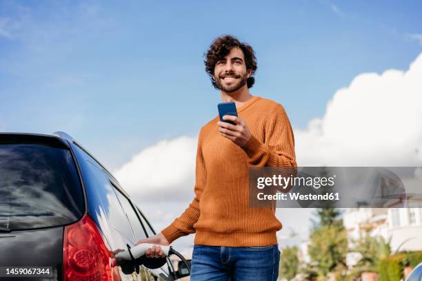 happy young man with mobile phone charging car at electric vehicle charging station - car mobile phone stock-fotos und bilder