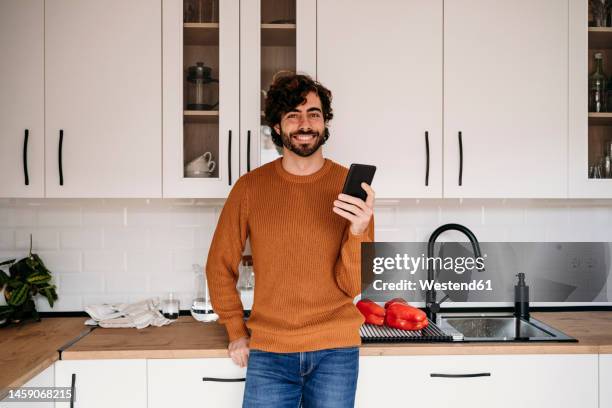 happy man with smart phone leaning on kitchen counter at home - homme cuisine photos et images de collection