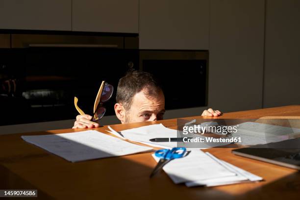 worried man hiding from financial bills on desk at home - hidden object stock pictures, royalty-free photos & images