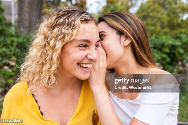 young woman whispering into friend's ear in park - ostördhet bildbanksfoton och bilder