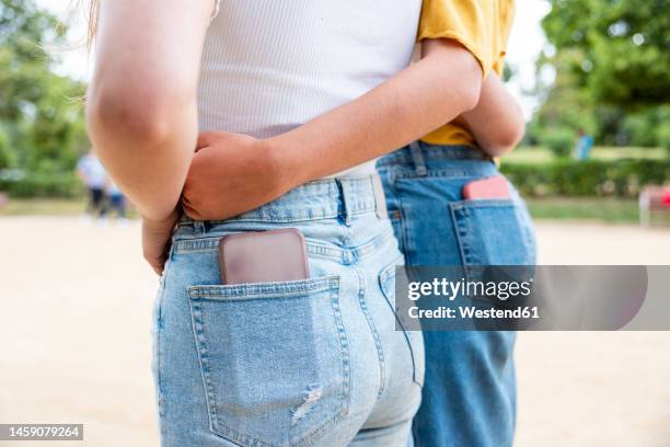 lesbian couple with smart phone in pockets embracing each other in park - jeans back stockfoto's en -beelden