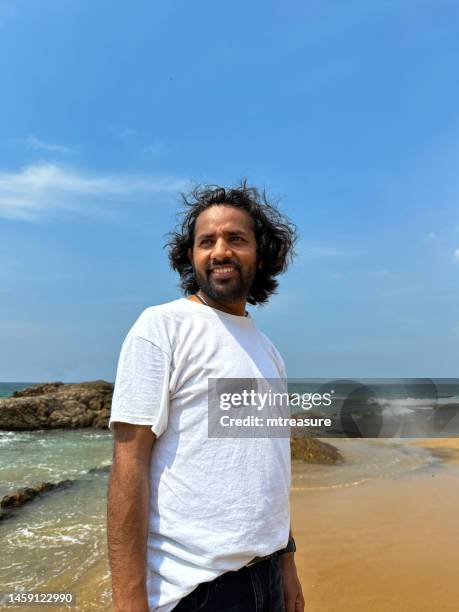 image of indian male tourist on holiday standing on beach looking at sea, sea tide coming in behind him, sandy beach, breaking waves, sunny blue sky, mount lavinia beach, colombia, sri lanka - tee srilanka stock pictures, royalty-free photos & images