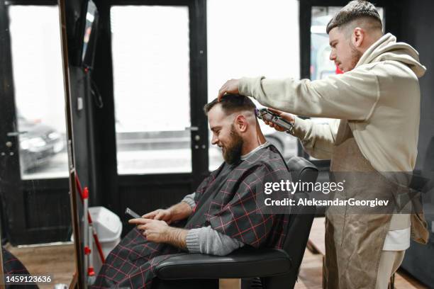 relaxed customer browsing haircut ideas while getting barber shop treatment - hair clipper stock pictures, royalty-free photos & images