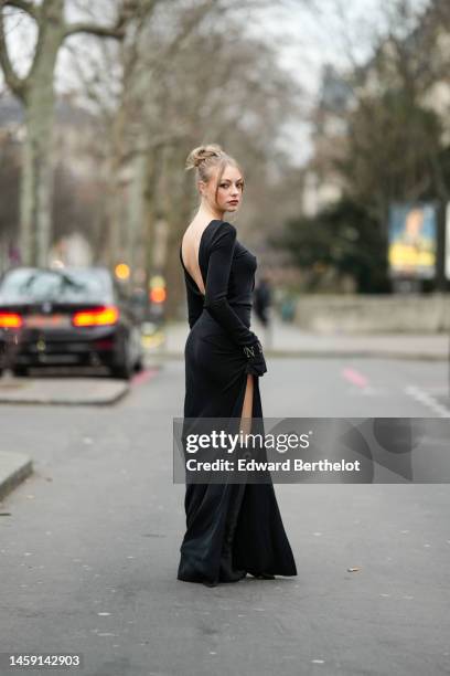 Anouchka Gauthier wears a black backless / long sleeves / slit long dress, a black feather handbag, black suede pointed heels knees boots / waders,...